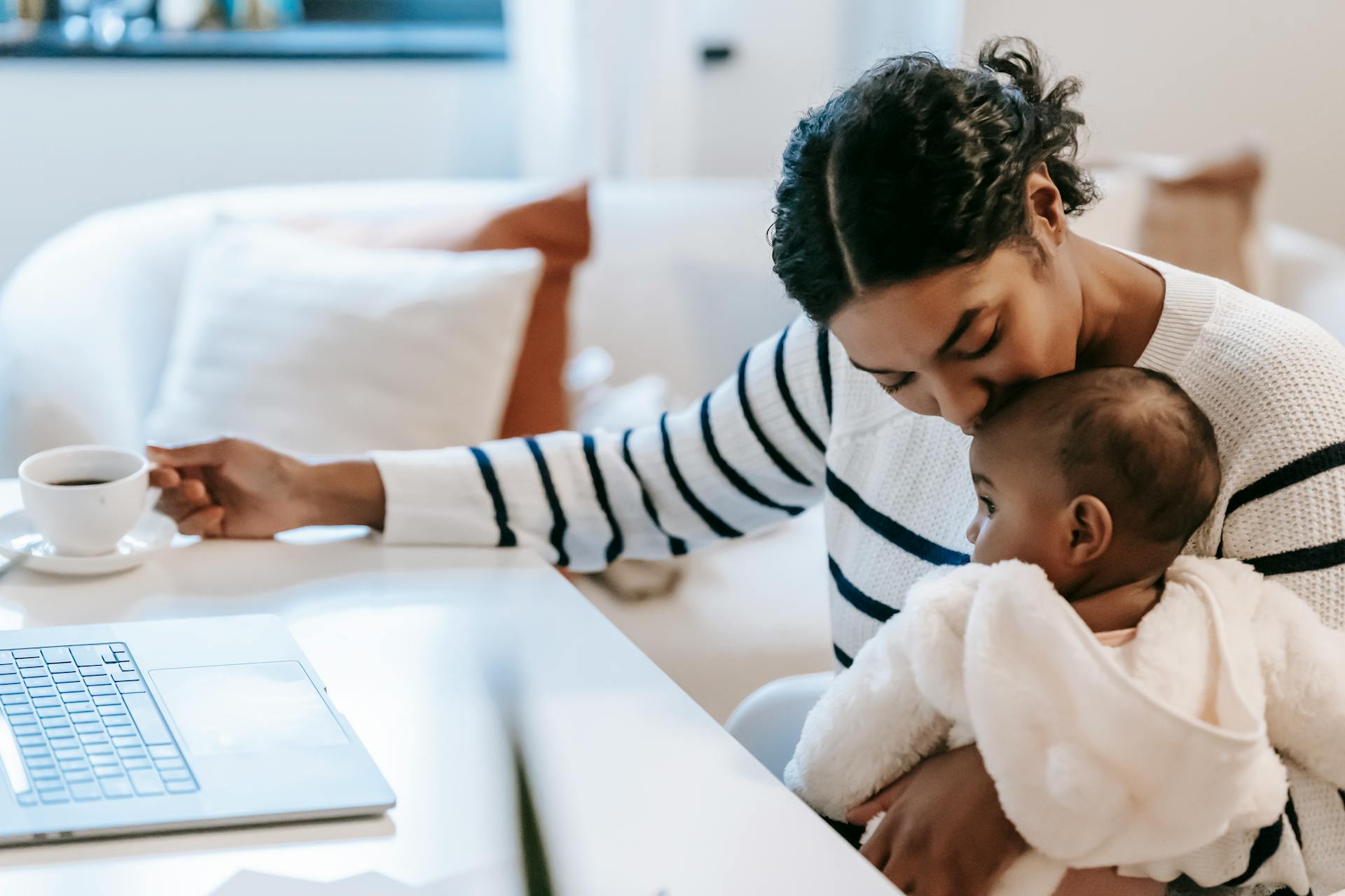 A working mother holds and kisses her baby on one arm while taking tea